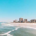 A imagem mostra a Praia da Barra da Tijuca, ensolarada com ondas suaves, cercada por prédios altos ao fundo. Algumas pessoas estão aproveitando o dia na areia e no mar, enquanto as montanhas aparecem ao longe sob um céu azul claro.