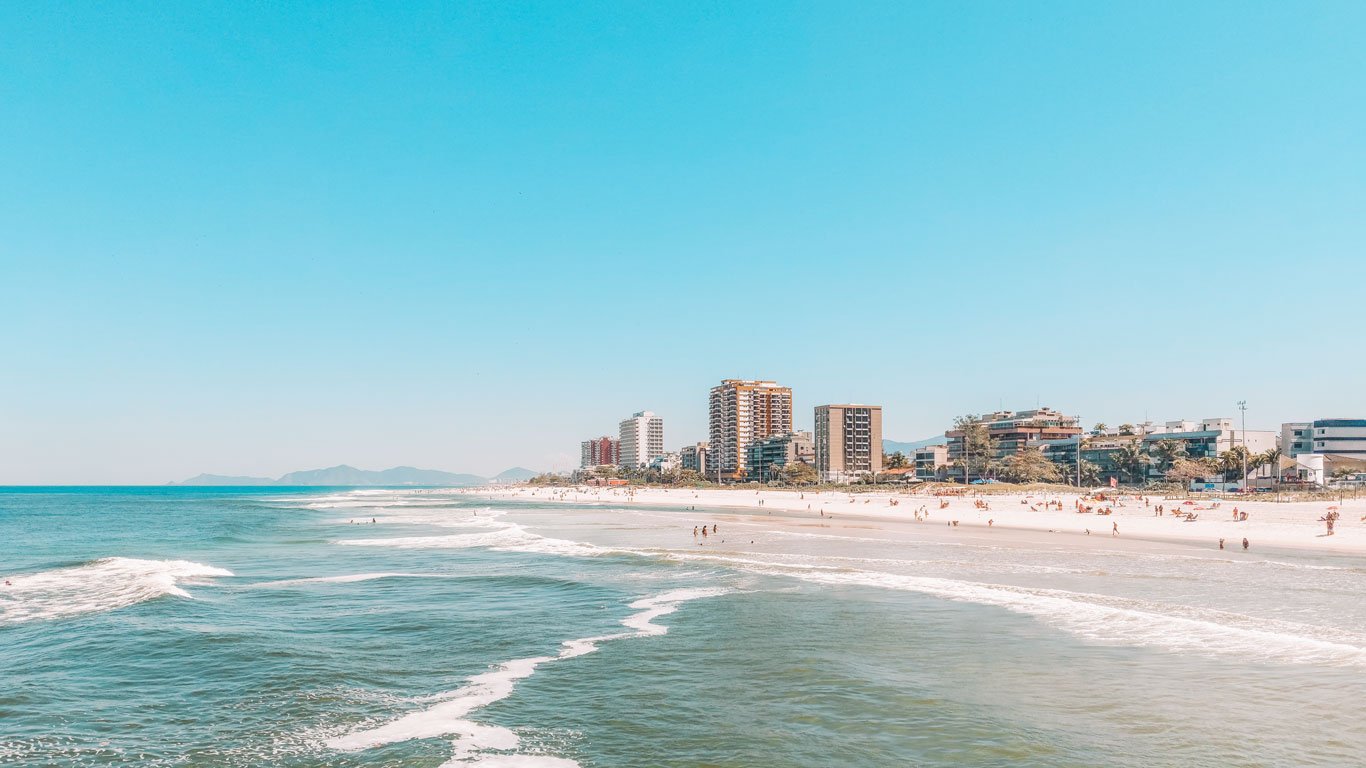 A imagem mostra a Praia da Barra da Tijuca, ensolarada com ondas suaves, cercada por prédios altos ao fundo. Algumas pessoas estão aproveitando o dia na areia e no mar, enquanto as montanhas aparecem ao longe sob um céu azul claro.