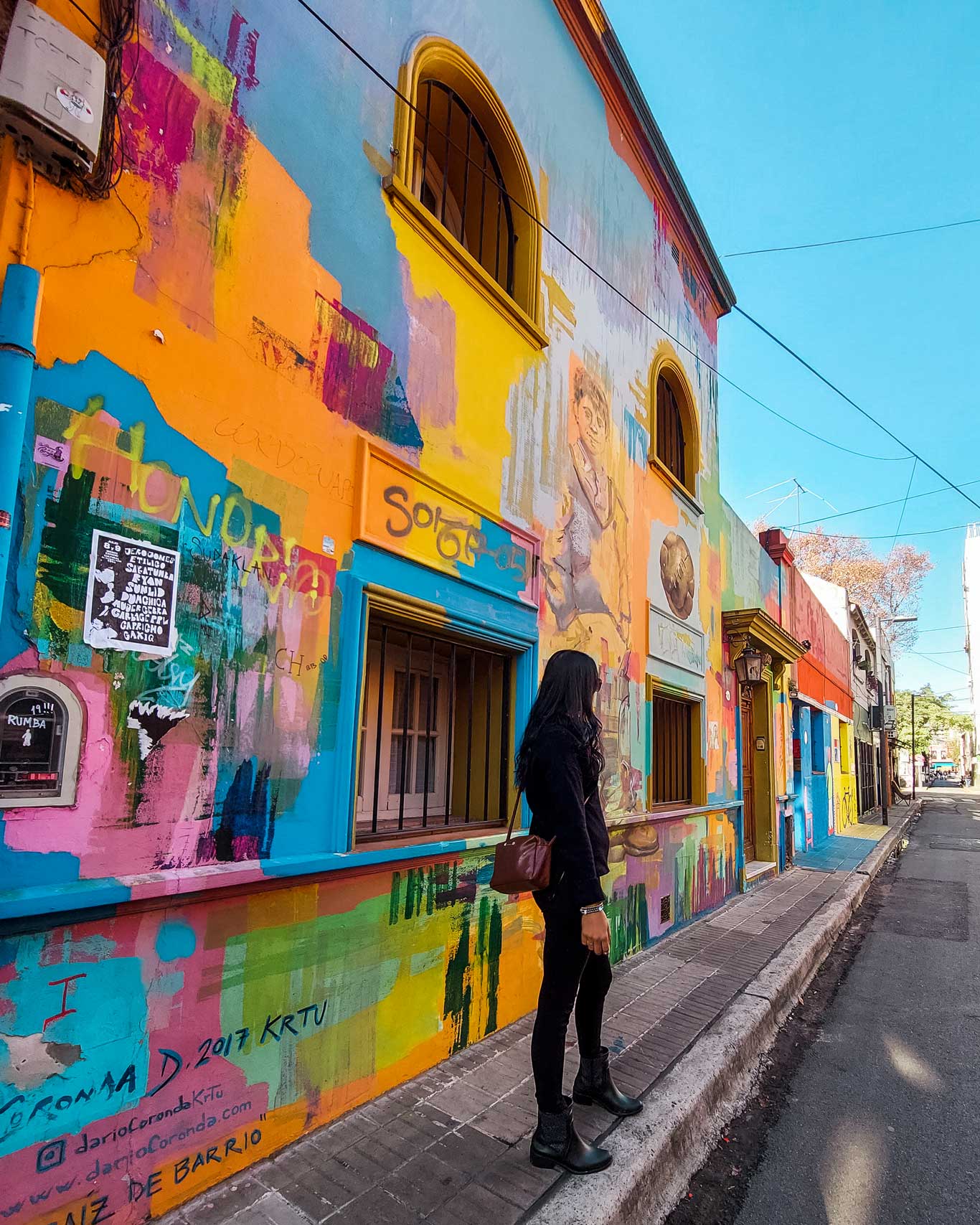 Mulher observando a fachada colorida de um edifício na Calle Russel, em Palermo, Buenos Aires. A parede é vibrante, com várias cores e grafites, incluindo ilustrações artísticas que cobrem toda a superfície. A mulher, vestida com roupas escuras, caminha pela calçada estreita, apreciando o ambiente artístico e dinâmico da rua.