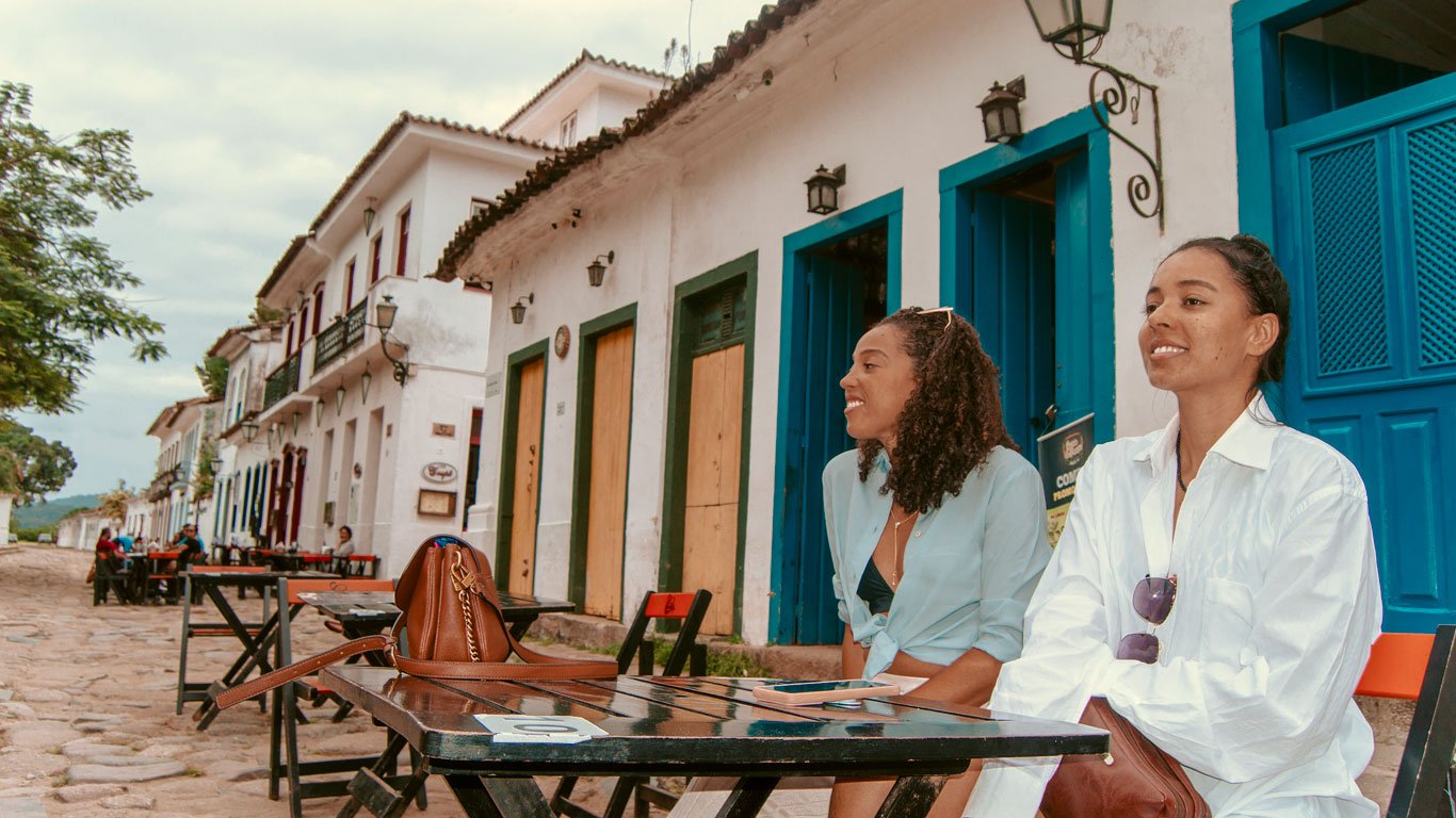 Duas mulheres sentadas em uma mesa ao ar livre em uma rua de paralelepípedos em Paraty, com casas históricas brancas e janelas de madeira coloridas ao fundo. Uma delas sorri, enquanto a outra parece estar observando o movimento.