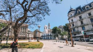 Museu do Cabildo em Buenos Aires, localizado na Plaza de Mayo. À esquerda, há uma árvore com galhos que se estendem sobre a calçada, e uma mulher caminhando com uma mochila vermelha. Ao fundo, vê-se a arquitetura histórica do Cabildo, com sua torre característica, e prédios antigos que cercam a praça sob um céu azul claro.