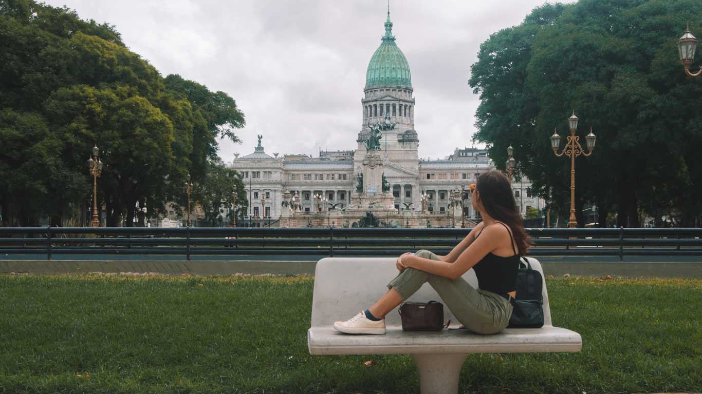 Mulher sentada em um banco na Plaza del Congreso, em Buenos Aires, contemplando o edifício do Congresso Nacional da Argentina ao fundo. O prédio imponente com sua cúpula verde se destaca entre as árvores e estátuas que adornam a praça.