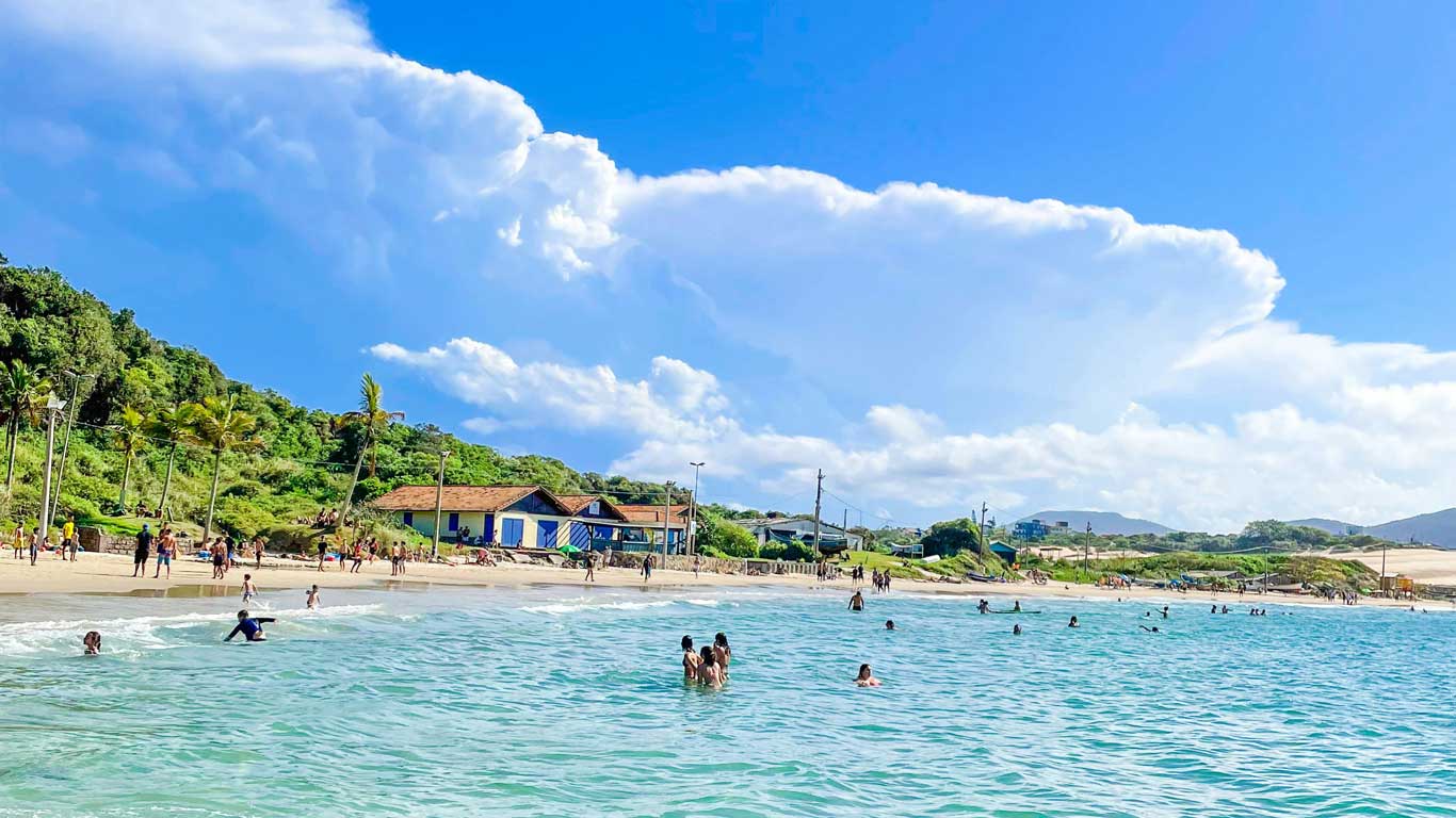 Praia dos Ingleses, com águas azuis claras, onde várias pessoas aproveitam o mar. Ao fundo, há casas cercadas pela vegetação tropical exuberante.