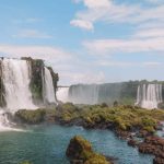 Cataratas do Iguaçu vista do lado brasileiro, mostrando várias quedas d'água caindo sobre penhascos rochosos cercados por vegetação verde e exuberante. A cena captura o fluxo poderoso da água com um céu claro ao fundo, destacando a beleza natural da paisagem.