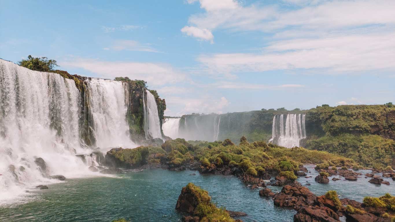 Cataratas do Iguaçu vista do lado brasileiro, mostrando várias quedas d'água caindo sobre penhascos rochosos cercados por vegetação verde e exuberante. A cena captura o fluxo poderoso da água com um céu claro ao fundo, destacando a beleza natural da paisagem.