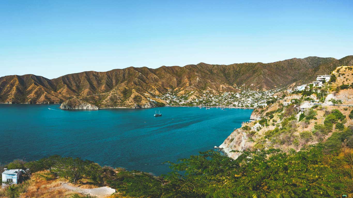 Uma baía tranquila de Taganga com águas azuis cercada por montanhas verdes. Pequenas embarcações são vistas ao longe, e a costa é pontilhada com casas e vegetação esparsa.