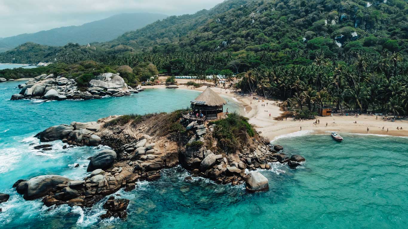 Vista aérea de uma pequena península rochosa no Parque Tayrona, com uma cabana no topo cercada por água cristalina. Ao fundo, uma praia de areia clara é envolta por vegetação densa e montanhas verdes.