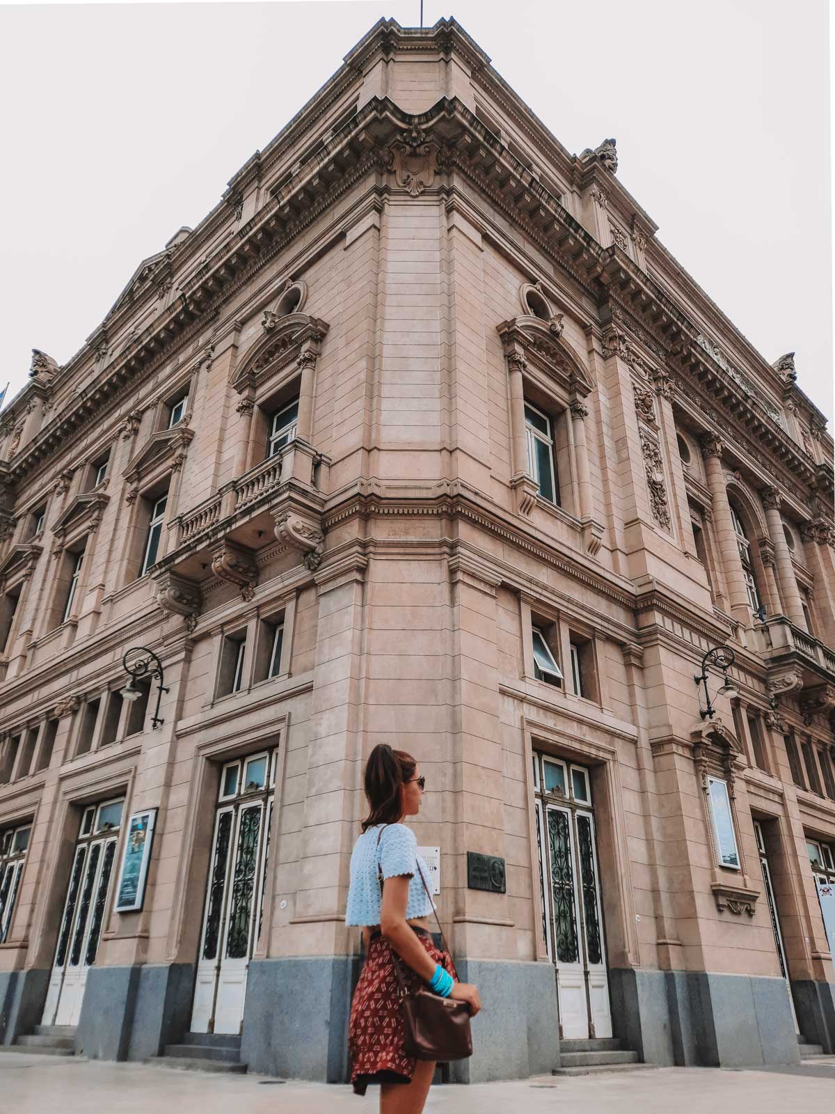 A imagem mostra uma mulher parada do lado de fora do Teatro Colón, em Buenos Aires, observando a fachada imponente do edifício. O teatro, com sua arquitetura clássica e detalhes ornamentados, destaca-se com suas janelas e molduras elegantes. A mulher veste uma blusa branca e uma saia estampada, segurando uma bolsa, enquanto aprecia a grandiosidade do prédio histórico.