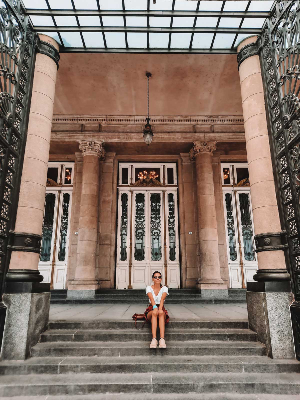 A imagem mostra a entrada do Teatro Colón em Buenos Aires, com uma mulher sentada nos degraus à frente. A estrutura é adornada por colunas clássicas e portões de ferro trabalhado, com portas altas ao fundo. O teto de vidro e as luminárias de época completam a imponência arquitetônica do local. A mulher está sorrindo e vestida casualmente, acrescentando uma sensação descontraída à cena.