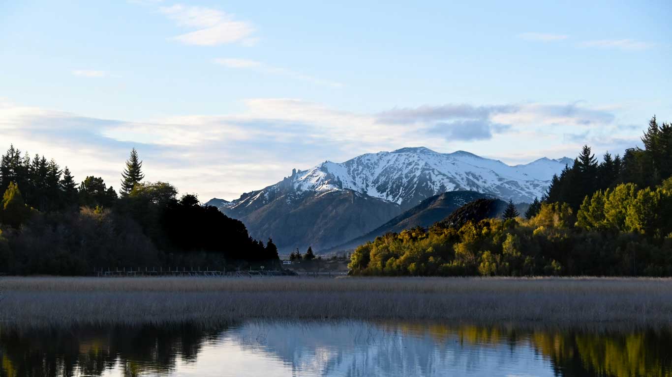 Paisagem de um lago de Bariloche refletindo montanhas nevadas ao fundo, com árvores densas nas margens e céu parcialmente nublado.