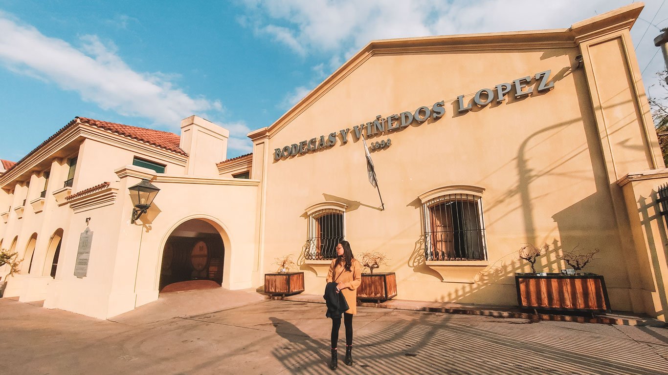 Mulher parada em frente à fachada da Bodega López, um prédio histórico com arquitetura em tons de bege e telhado de telhas vermelhas, localizado sob um céu azul. A entrada da vinícola exibe o nome "Bodegas y Viñedos López" e o ano de fundação "1898", destacando a tradição e elegância do local, ideal para quem aprecia enoturismo em Mendoza.