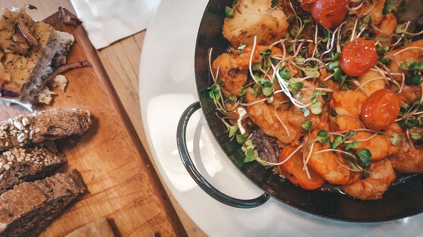 Prato colorido de frutos do mar servido em uma panela de ferro, decorado com microverdes e tomates-cereja, ao lado de uma tábua com fatias de pão rústico, incluindo opções com sementes e focaccia. A apresentação combina sabores frescos e texturas variadas, perfeita para harmonizar com vinhos em um ambiente de vinícola.