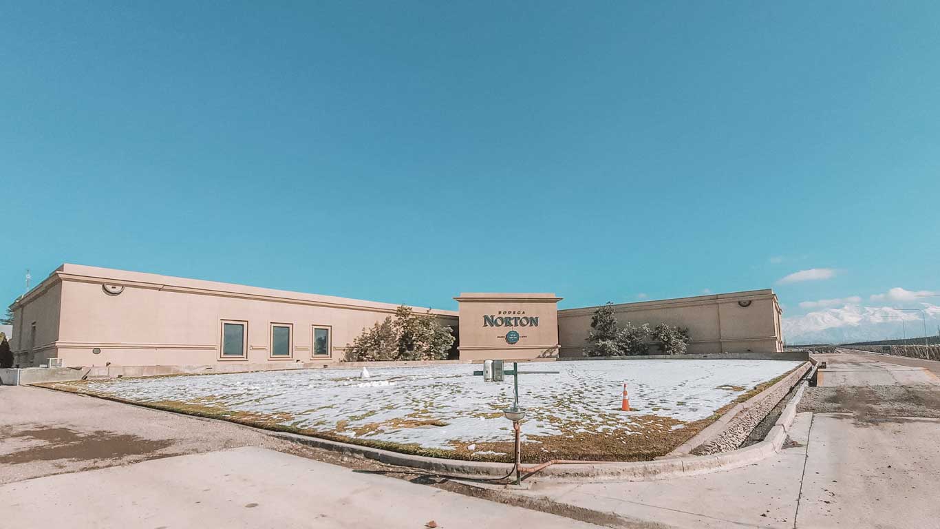 Fachada da Bodega Norton em Mendoza, Argentina, em um dia de céu claro. O edifício apresenta arquitetura simples e moderna, com o nome "Bodega Norton" destacado no centro. No chão, uma fina camada de neve cobre o gramado, e ao fundo, é possível ver as montanhas dos Andes, criando um cenário pitoresco e convidativo para visitantes de vinícolas.