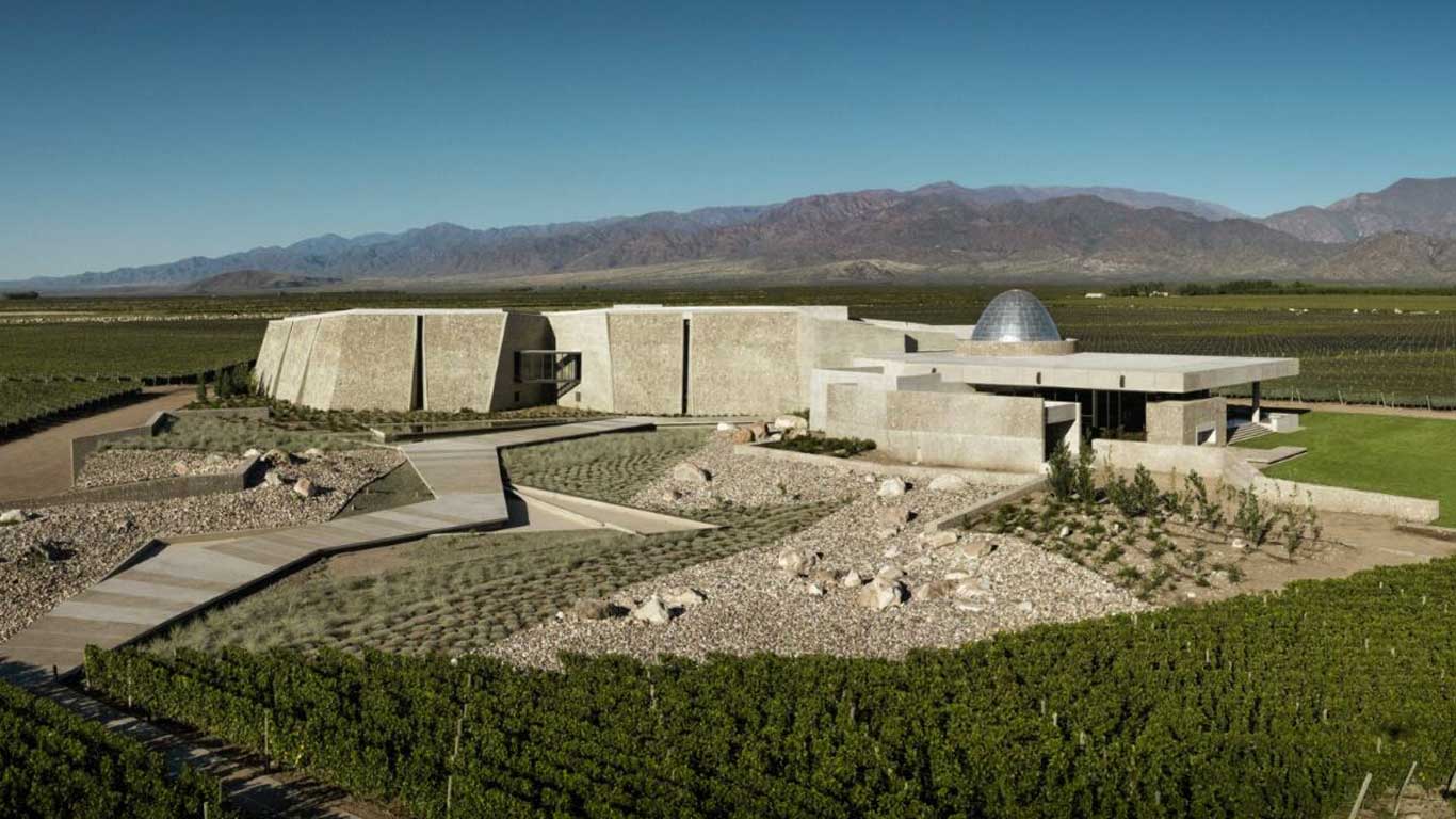 A Bodega Zuccardi, uma das melhores vinícolas de Mendoza, possui uma arquitetura moderna e impressionante, com formas geométricas em pedra que se integram ao ambiente natural dos vinhedos ao redor. Ao fundo, a imponente cordilheira dos Andes completa a paisagem, reforçando o ambiente único e sofisticado que caracteriza esta vinícola renomada, conhecida pela inovação e sustentabilidade.