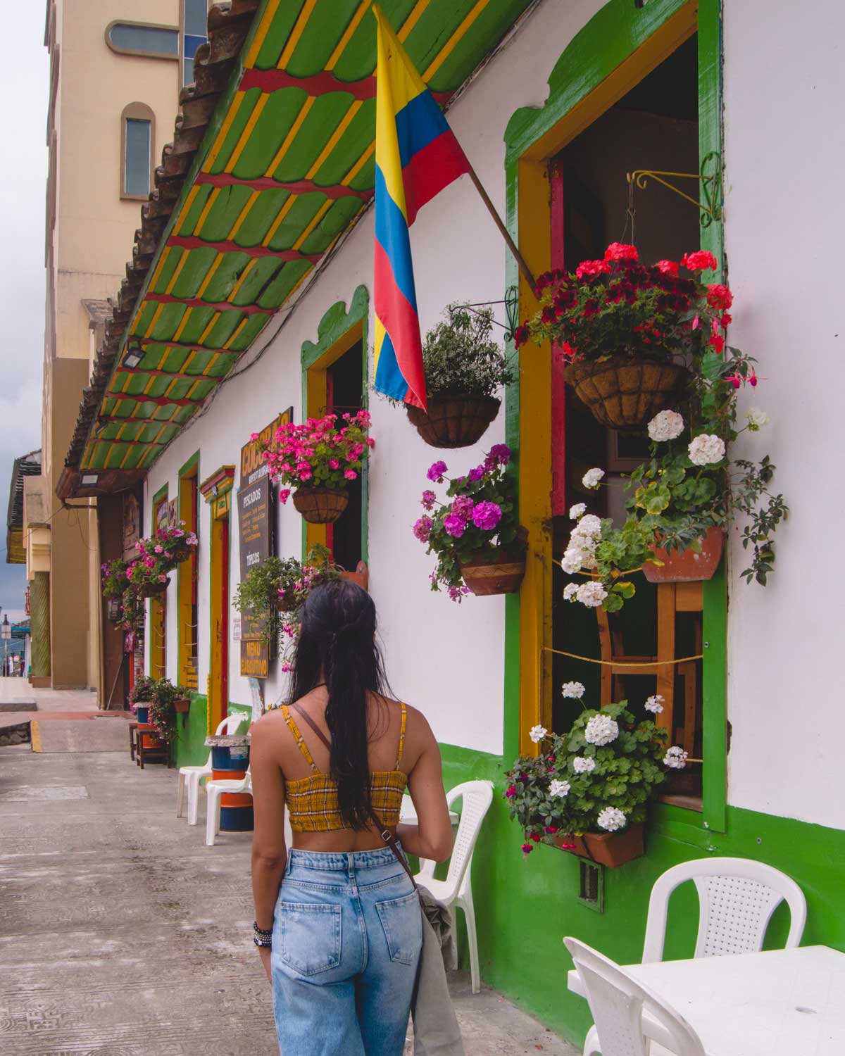 Uma mulher de costas caminha pelo centro de Salento, o melhor lugar onde ficar em Salento na Colômbia, em frente a um prédio decorado com cores vivas. A fachada é branca com detalhes verdes, e há vasos de flores pendurados nas janelas. A bandeira da Colômbia está hasteada ao lado da porta.