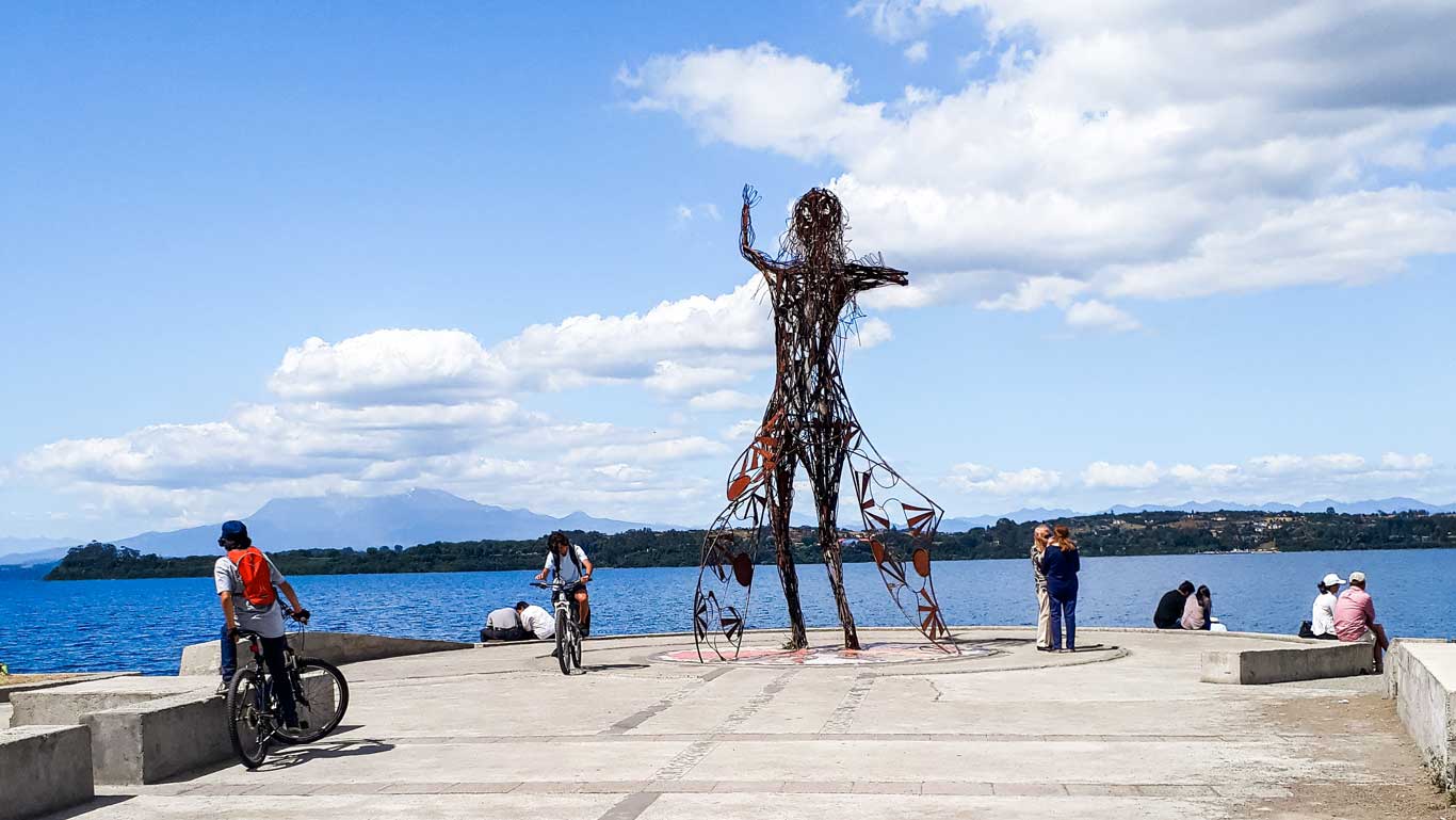 Imagem da Princesa Licarayén, uma escultura de metal estilizada de uma figura humana com os braços levantados, instalada próximo ao Lago Llanquihue, uma das melhores opções de o que fazer em Puerto Varas. Ao redor, ciclistas e visitantes apreciam o local com o cenário montanhoso ao fundo.