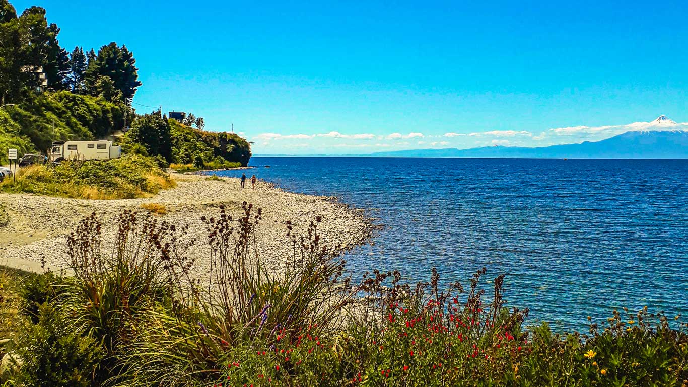 Uma praia com pequenas pedras e vegetação rasteira nas margens do Lago Llanquihue, em Puerto Varas, ao fundo vulcões cobertos de neve no horizonte. Há pessoas caminhando à beira d'água, aproveitando o clima ensolarado.