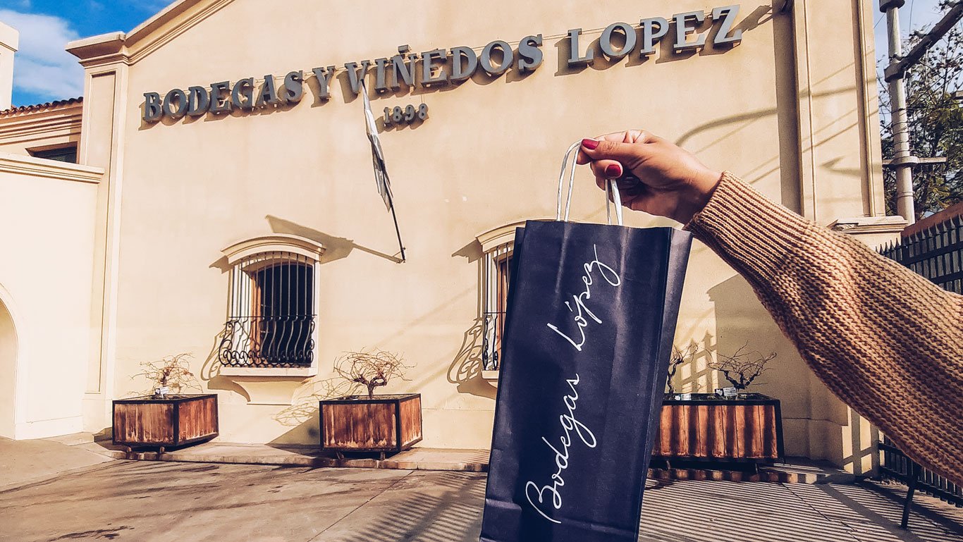 Mão segurando uma sacola preta elegante com o logotipo da Bodega López em destaque, em frente à fachada da vinícola em Mendoza, que exibe o nome "Bodegas y Viñedos López" e o ano "1898". A imagem transmite a experiência de uma compra especial de vinhos, combinando tradição e sofisticação.