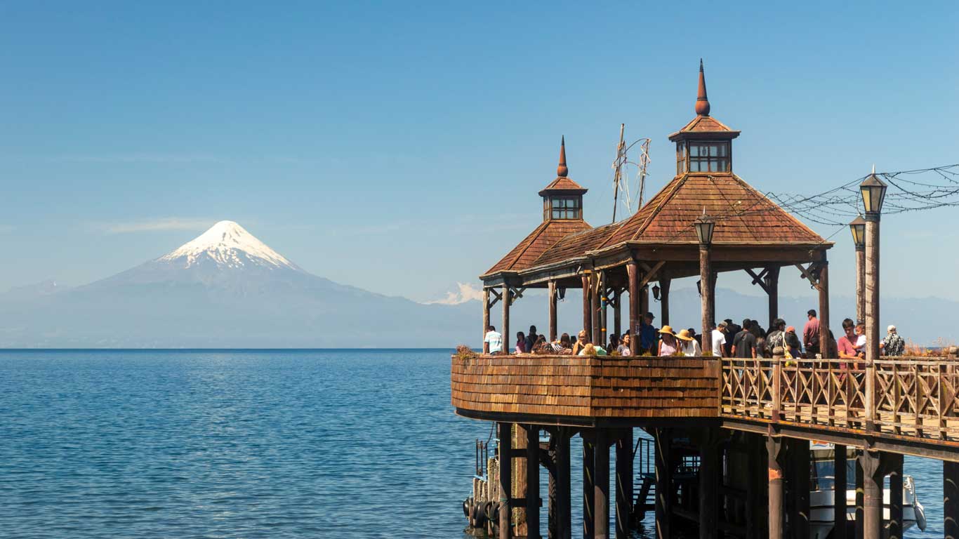 Um pavilhão de madeira sobre o Lago Llanquihue em Frutillar, com visitantes apreciando a vista do Vulcão Osorno ao fundo. A construção rústica é cercada por água calma e o céu aberto.