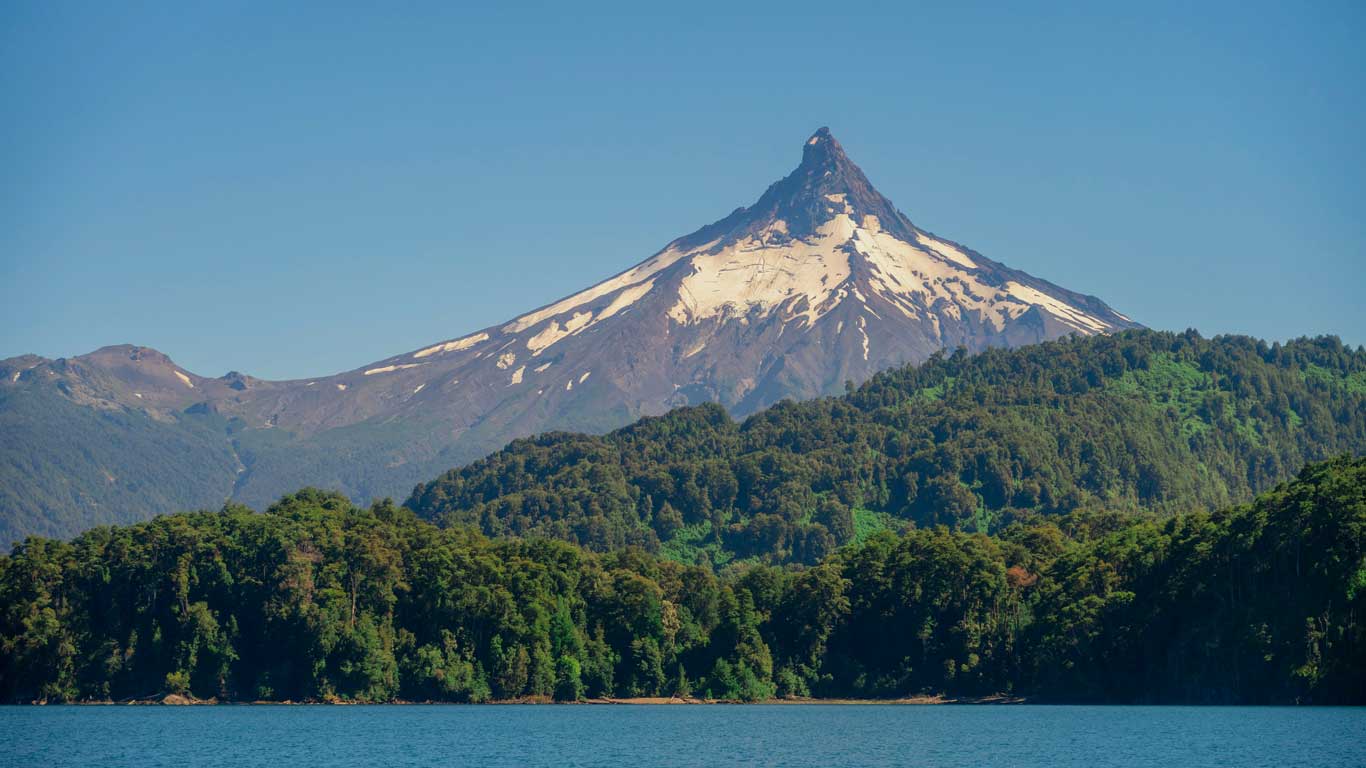 Vista panorâmica do Lago de Todos los Santos, com montanhas e vegetação exuberante ao redor. O dia ensolarado realça o azul intenso do lago.