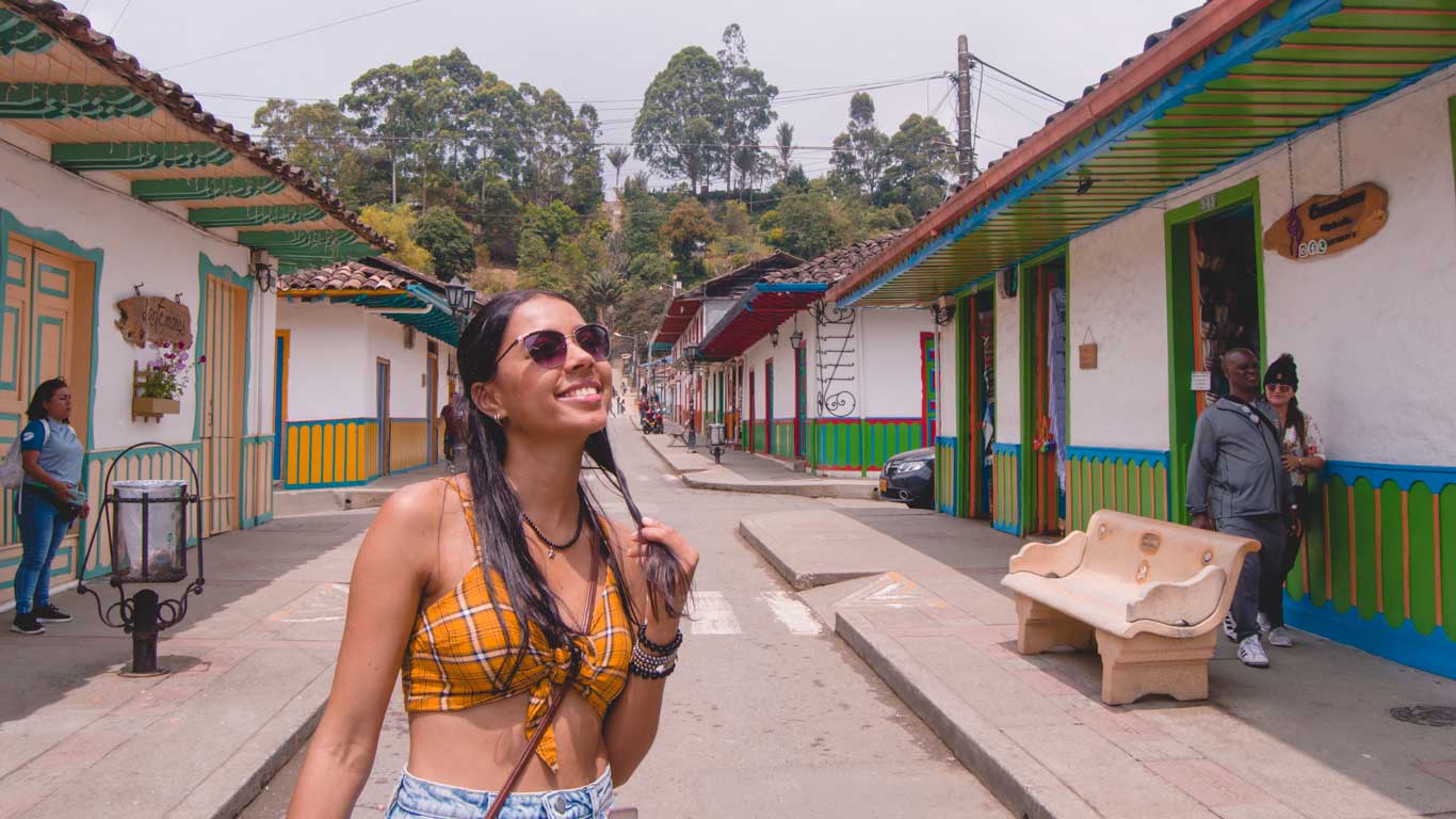 Uma jovem sorri enquanto caminha por uma rua de Salento, cercada por construções coloridas com detalhes verdes, amarelos e azuis nas fachadas. Ao fundo, há vegetação e outras pessoas explorando o local.