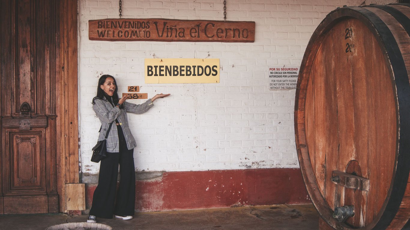 Mulher sorridente apontando para uma placa com a palavra "BIENBEBIDOS" na entrada da Viña el Cerno, em Mendoza. A cena inclui elementos rústicos, como uma grande porta de madeira e um barril de vinho, criando uma atmosfera acolhedora e divertida que celebra o espírito da degustação de vinhos.