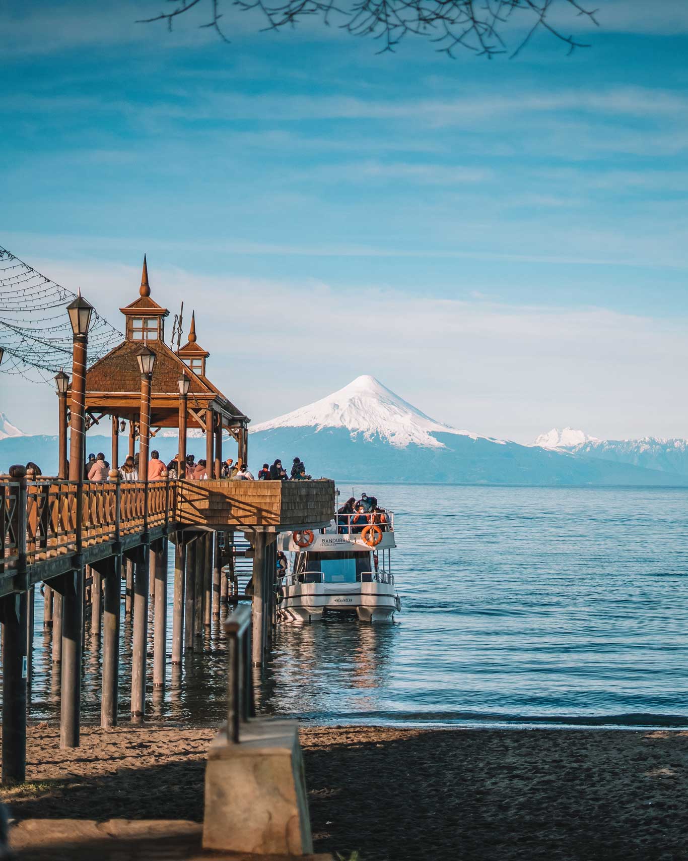 Imagem da cidade de Frutillar, no Chile, situada à beira do Lago Llanquihue. Em destaque, um pier de madeira com visitantes apreciando a paisagem, enquanto um barco está atracado. Ao fundo, o majestoso Vulcão Osorno com seu cume coberto de neve completa o cenário natural deslumbrante. Select 54 more words to run Humanizer.