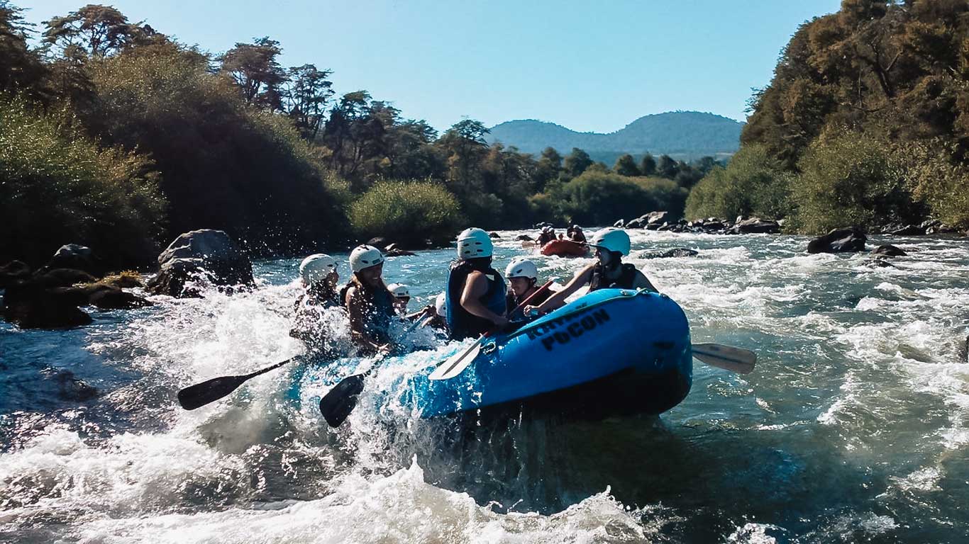 Um grupo de pessoas em um bote azul enfrentando as corredeiras de um rio em meio à vegetação densa em um passeio de rafting em Pucón, no Chile. Todos estão usando capacetes e coletes salva-vidas enquanto remam pelas águas agitadas.