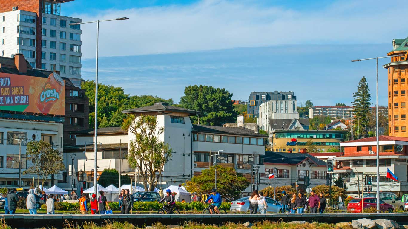 Paisagem urbana com prédios modernos e históricos, com destaque para uma igreja de torre alta no centro de Puerto Montt. Há um movimento de pessoas e carros pela área, e outdoors publicitários decoram alguns edifícios.