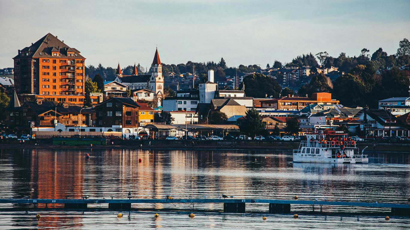 Vista de Puerto Varas a partir do lago, com prédios coloridos e uma igreja ao fundo. Uma embarcação branca navega pela água calma ao entardecer, refletindo o céu e a cidade na superfície do Lago Llanquihue.