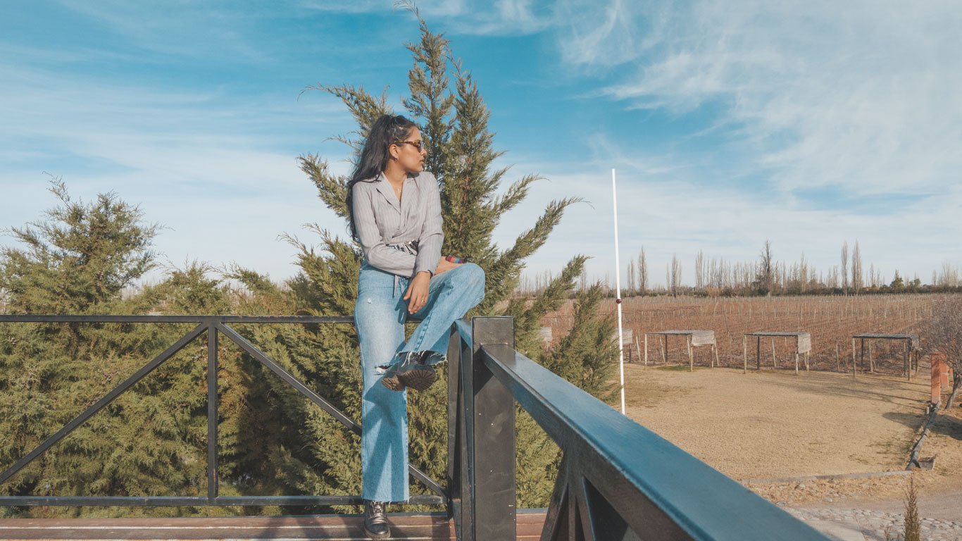 Mulher sentada em uma cerca de metal, usando óculos de sol, camisa listrada e calça jeans, olhando para o lado em um ambiente de vinhedo. Ao fundo, árvores verdes e uma paisagem ampla de vinhas sob um céu azul claro, criando uma atmosfera tranquila e serena.