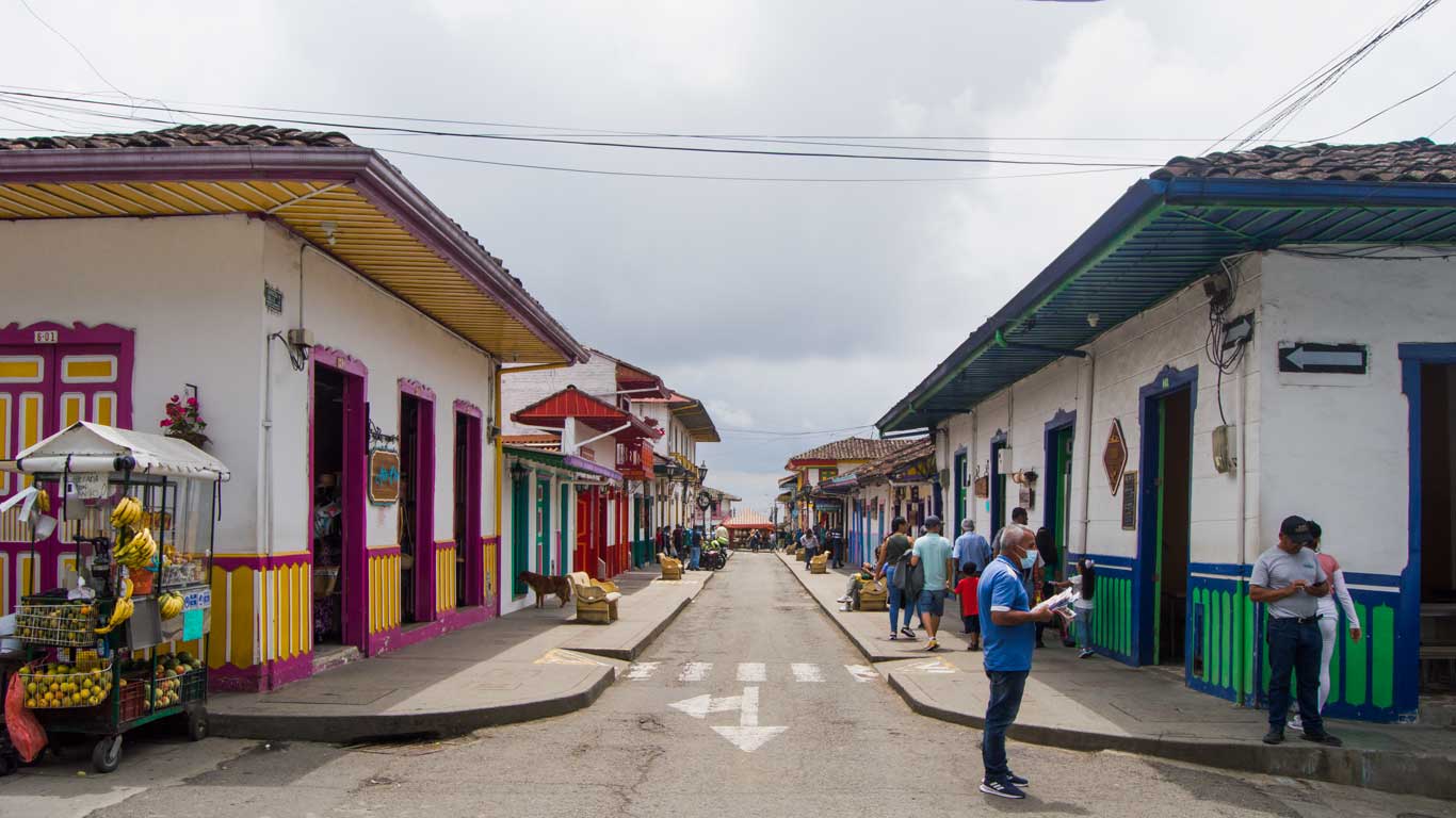 Rua de Salento movimentada, com construções coloridas e detalhes nas portas e janelas em verde, azul e amarelo. Uma barraca vende frutas, e algumas pessoas caminham e observam a paisagem urbana.