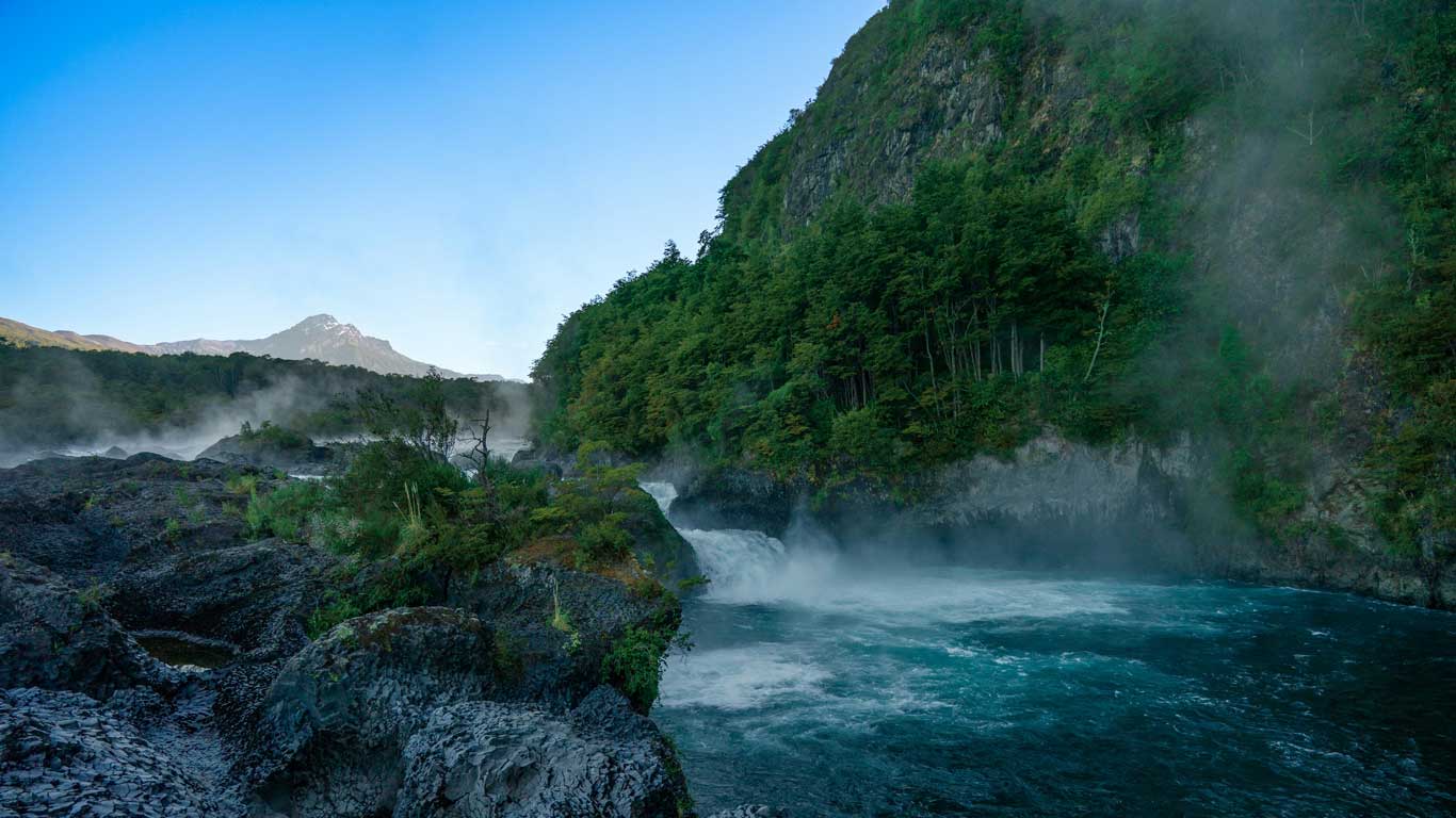 Imagem das cachoeiras de Petrohué em um ambiente montanhoso e cercado por mata verde. A névoa e o movimento da água criam uma atmosfera de tranquilidade, enquanto o céu azul é visível ao fundo.