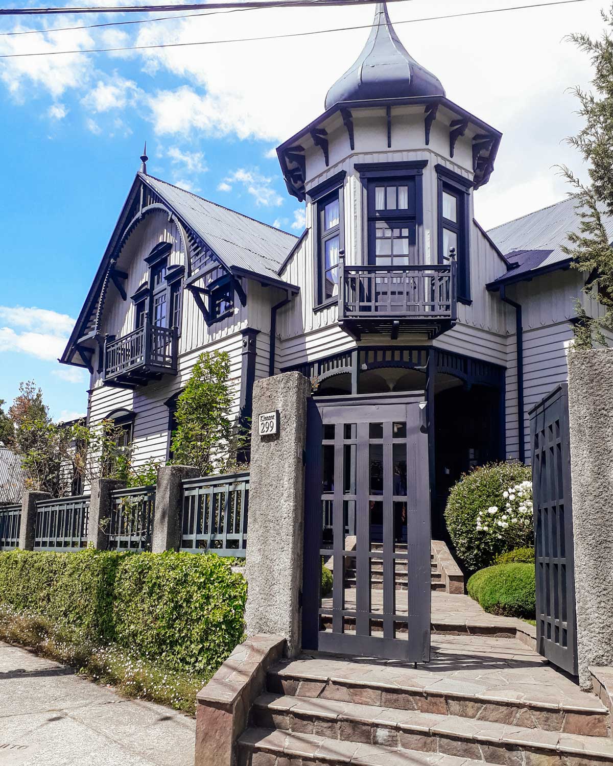 Uma casa tradicional de estilo arquitetônico alemão em Puerto Varas, Chile, com paredes brancas, detalhes em preto e telhado inclinado com uma torre distinta. O jardim bem cuidado rodeia a propriedade, com arbustos verdes e flores brancas. O céu limpo e a fachada elegante realçam a beleza desta construção típica da região.