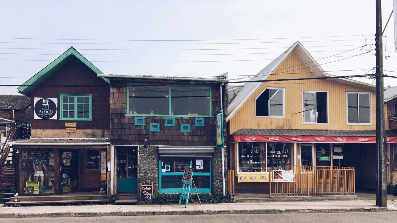 Rua comercial em Dalcahue, na ilha de Chiloé, Chile. A imagem apresenta dois edifícios lado a lado: à esquerda, um restaurante com fachada de madeira escura e janelas verde-água; à direita, uma loja amarela com varanda cercada e toldo vermelho com a logo da Coca-Cola. O cenário é complementado por fios elétricos no alto e uma rua asfaltada à frente.