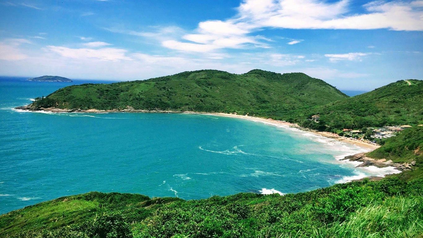 Vista aérea da Praia Brava em Búzios, mostrando uma enseada de águas azul-turquesa cercada por colinas verdes cobertas de vegetação. A praia de areia clara se estende ao longo da costa, com ondas suaves tocando a margem e algumas construções à direita. O céu azul com nuvens brancas completa o cenário paradisíaco.