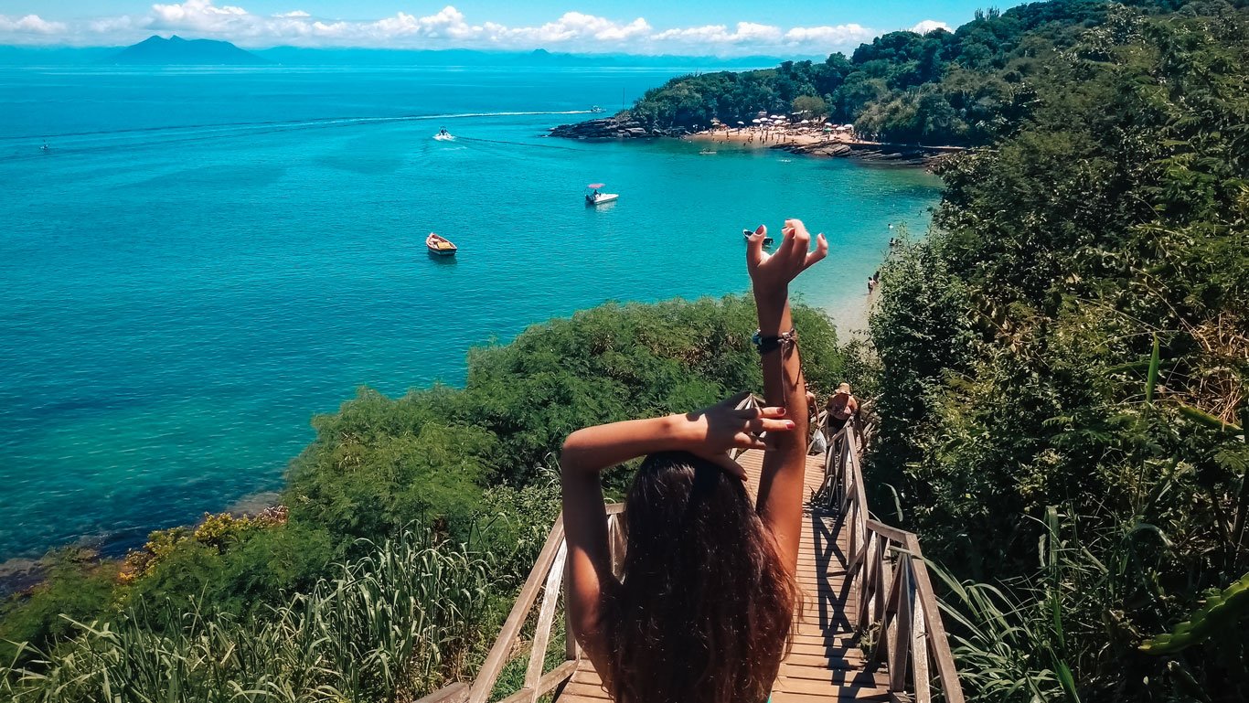 Uma pessoa de cabelos longos e braços levantados desce uma escada de madeira cercada por vegetação em direção às águas turquesas da Praia Azeda, uma das melhores praias de Búzios, Brasil. Pequenos barcos flutuam no mar calmo, com uma praia de areia e uma floresta densa ao fundo. O céu está claro e azul, com montanhas distantes no horizonte.