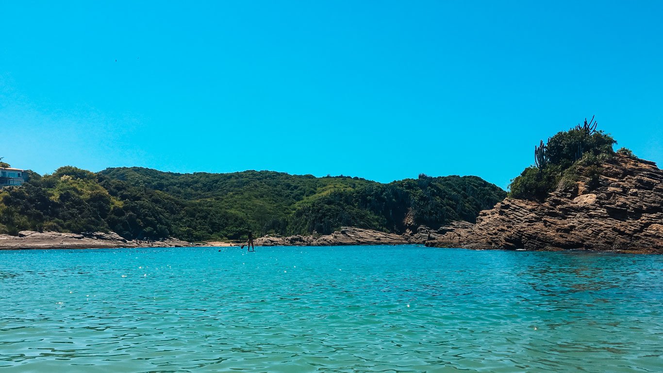 Vista da Praia da Ferradurinha em Búzios, com águas cristalinas e tranquilas em primeiro plano. Ao fundo, há colinas cobertas por vegetação e formações rochosas à direita, sob um céu azul sem nuvens. Algumas pessoas estão aproveitando o mar.