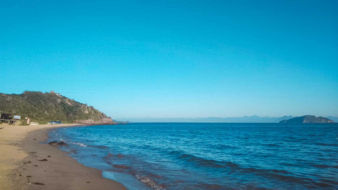 Praia da Tartaruga em Búzios, com uma faixa de areia dourada à esquerda e o mar azul estendendo-se à direita. Ao fundo, uma colina verde com vegetação, além de pequenas ilhas visíveis no horizonte sob um céu claro e azul.