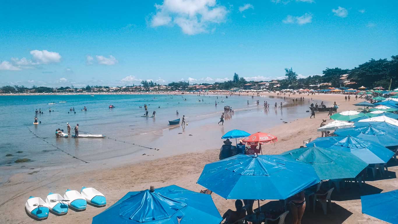 Praia de Geribá em Búzios, com muitas pessoas aproveitando o mar e a areia sob um céu azul. Fileiras de guarda-sóis coloridos, predominantemente azuis, cobrem a área à direita da imagem, enquanto caiaques estão alinhadas na areia. No fundo, casas e árvores complementam a paisagem litorânea movimentada.