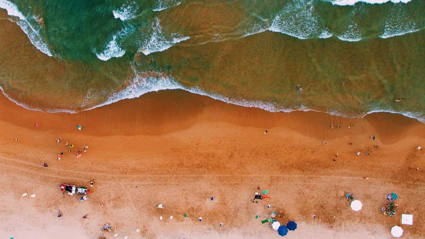 Vista aérea da Praia do Forno em Búzios, com ondas do mar verde-esmeralda quebrando suavemente na areia avermelhada. Pessoas estão espalhadas pela praia, algumas sob guarda-sóis e outras brincando na beira da água.