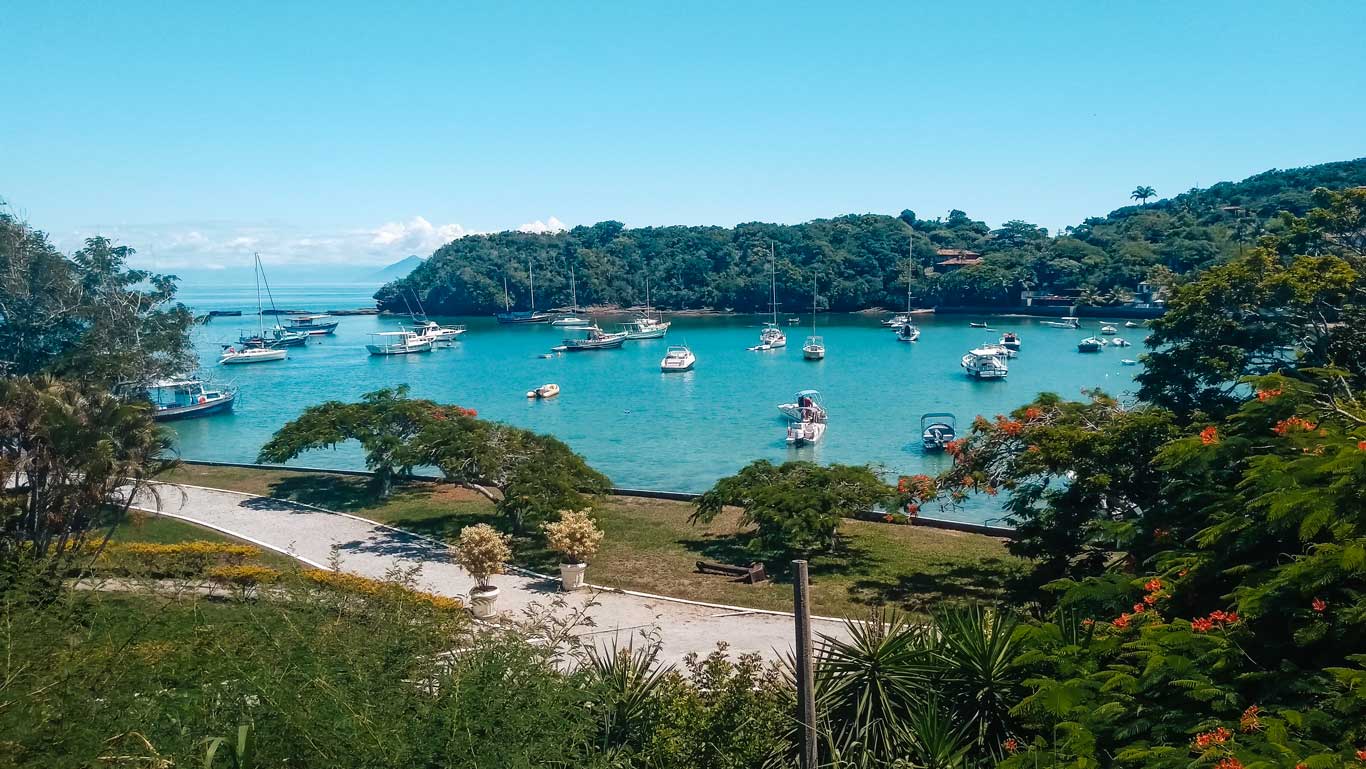 Praia dos Ossos em Búzios, com vários barcos ancorados em águas calmas e azul-turquesa. Em primeiro plano, árvores e flores emolduram a paisagem, enquanto uma trilha pavimentada passa pelo jardim à beira-mar. Ao fundo, colinas verdes e casas completam a vista sob um céu azul claro.