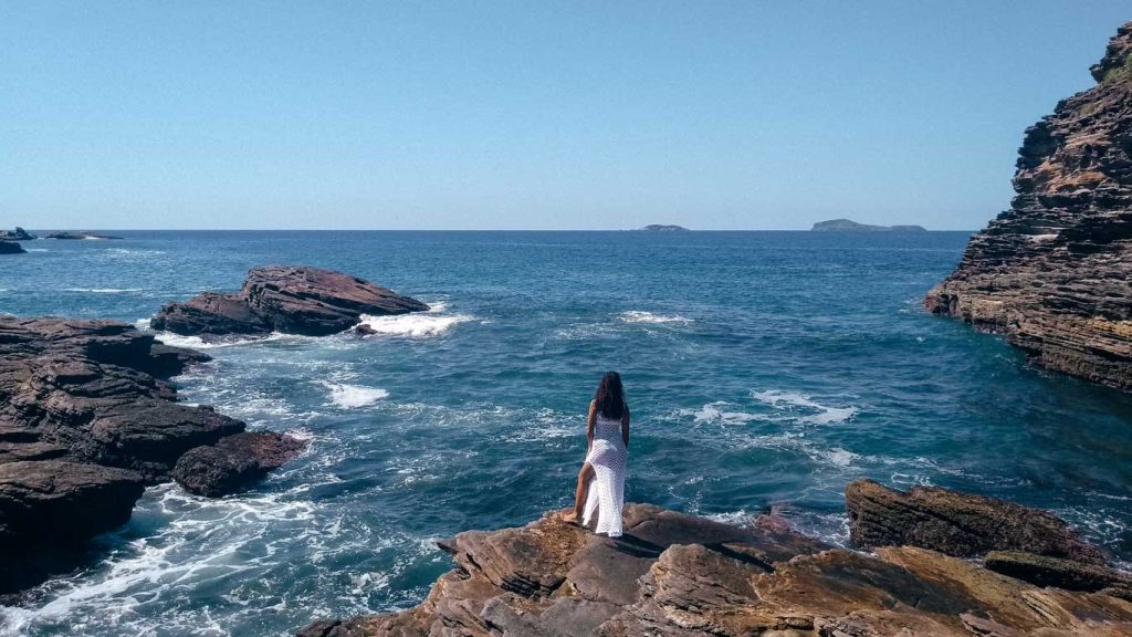 Uma mulher com um vestido branco longo está em pé sobre rochas à beira-mar, olhando para o vasto oceano azul com pequenas ondas quebrando nas pedras. A cena mostra formações rochosas escuras, um céu claro e ilhas distantes no horizonte.