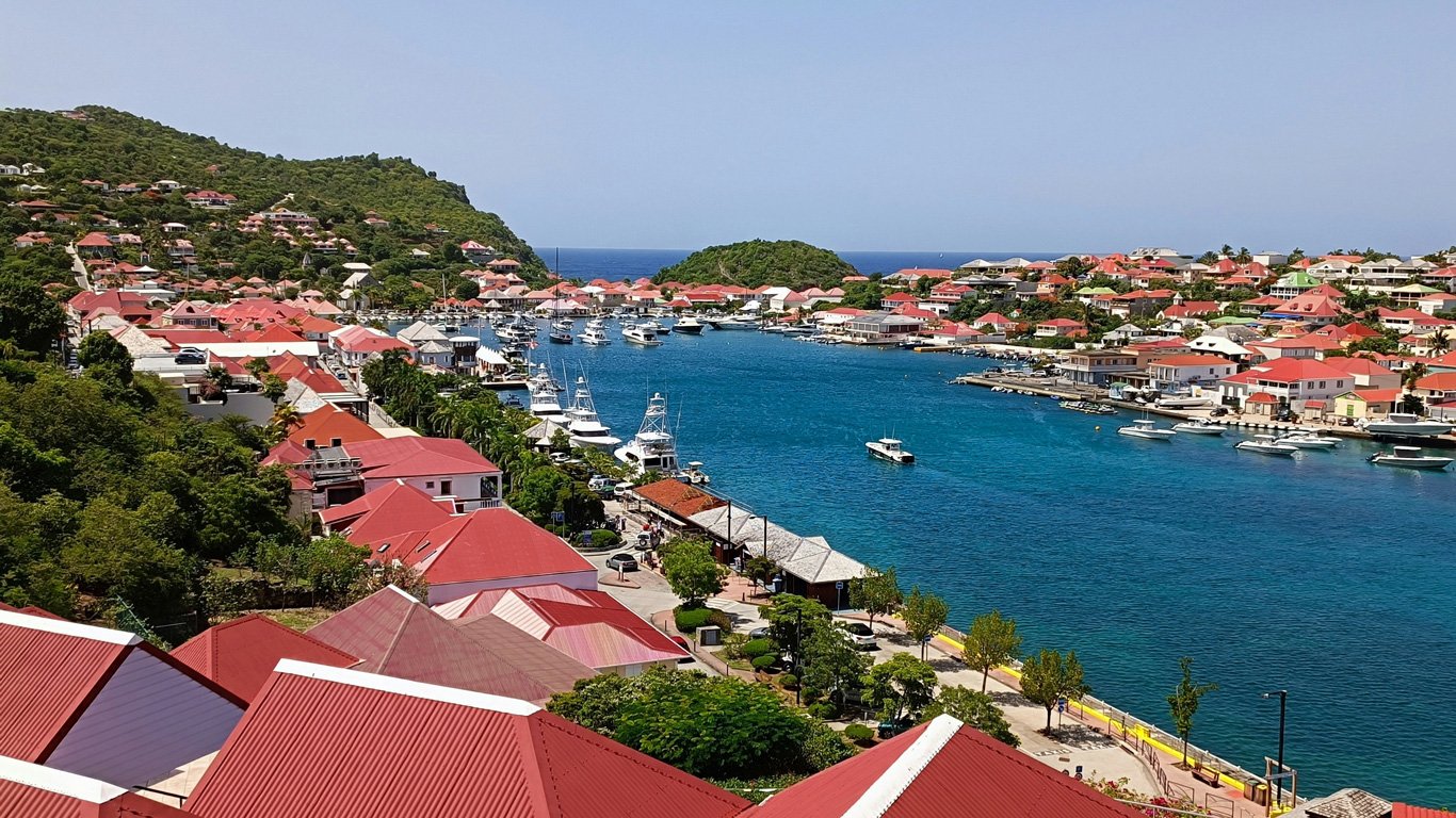 Uma vista panorâmica da cidade costeira de Saint-Barthélemy, com telhados vermelhos característicos contrastando com o azul intenso do mar. O porto está repleto de iates e barcos ancorados, enquanto as ruas arborizadas e bem cuidadas acompanham a orla. No fundo, colinas verdes cercam a cidade, criando um cenário tropical sofisticado e charmoso.