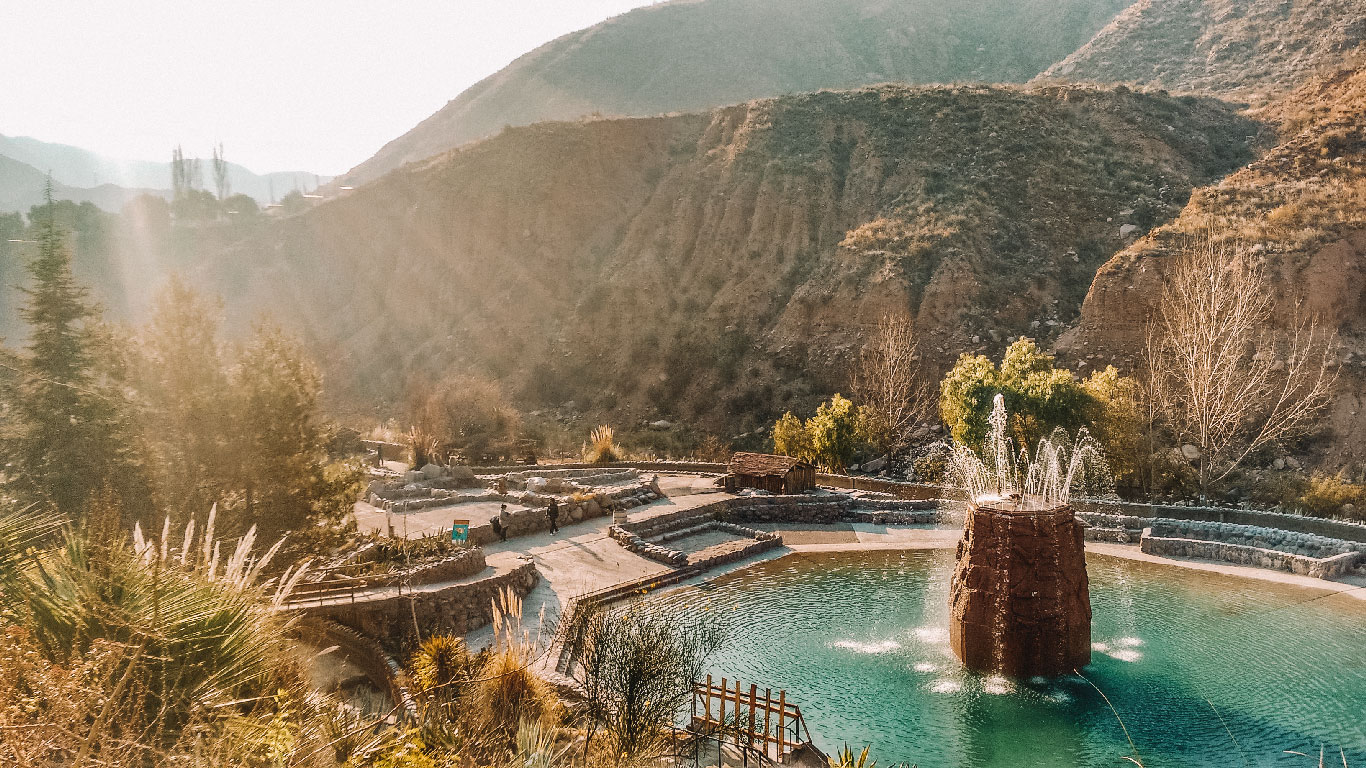 Imagem de um cenário nas Termas de Cacheuta, em Mendoza, Argentina, mostrando uma piscina termal com uma fonte central cercada por montanhas áridas e vegetação típica da região. O sol ilumina o local, criando um efeito dourado que destaca a beleza natural e a tranquilidade da área. Ao redor da piscina, há caminhos pavimentados e pequenas construções rústicas, compondo um ambiente relaxante e acolhedor.