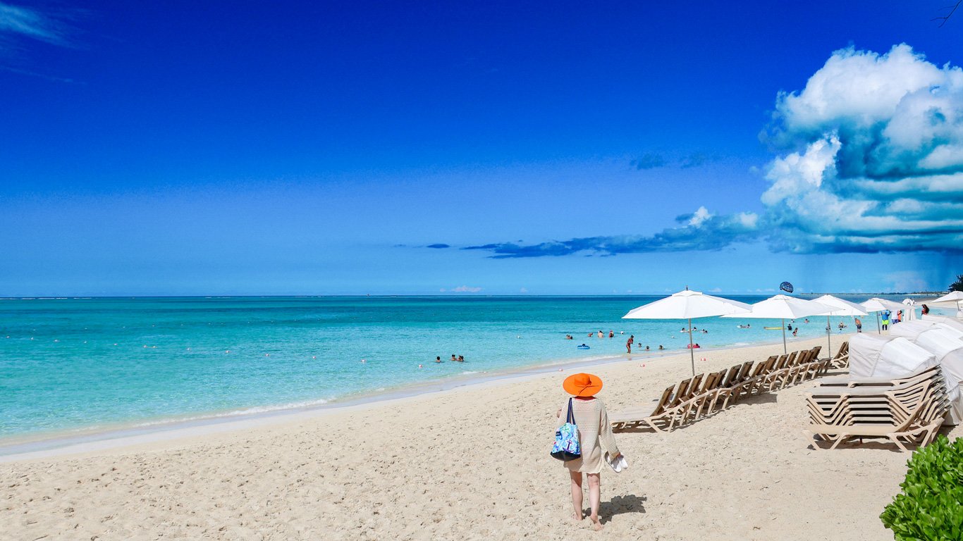 Uma praia paradisíaca em Turks and Caicos, com areia branca e águas azul-turquesa cristalinas. Uma mulher, vestindo um chapéu laranja e carregando uma bolsa de praia, caminha descalça na areia em direção ao mar. Espreguiçadeiras alinhadas sob guarda-sóis brancos contrastam com o céu azul intenso, onde nuvens dramáticas se formam ao fundo.