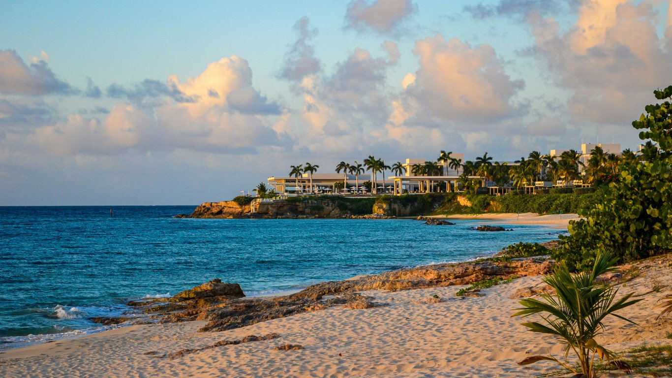 Uma praia de areia dourada em Anguilla, com águas cristalinas e um o resort Four Seasons Anguilla ao fundo, rodeado por palmeiras e falésias, sob um céu azul com nuvens suaves.