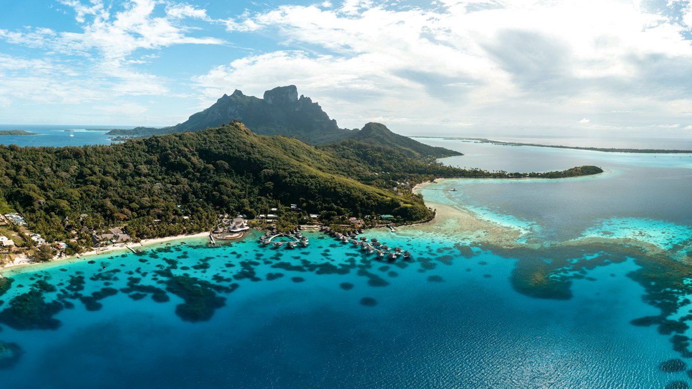 Uma vista aérea de Bora Bora com o monte Otemanu ao fundo, rodeado por lagoas azul-turquesa e bangalôs sobre a água, um destino de sonho no Pacífico Sul.