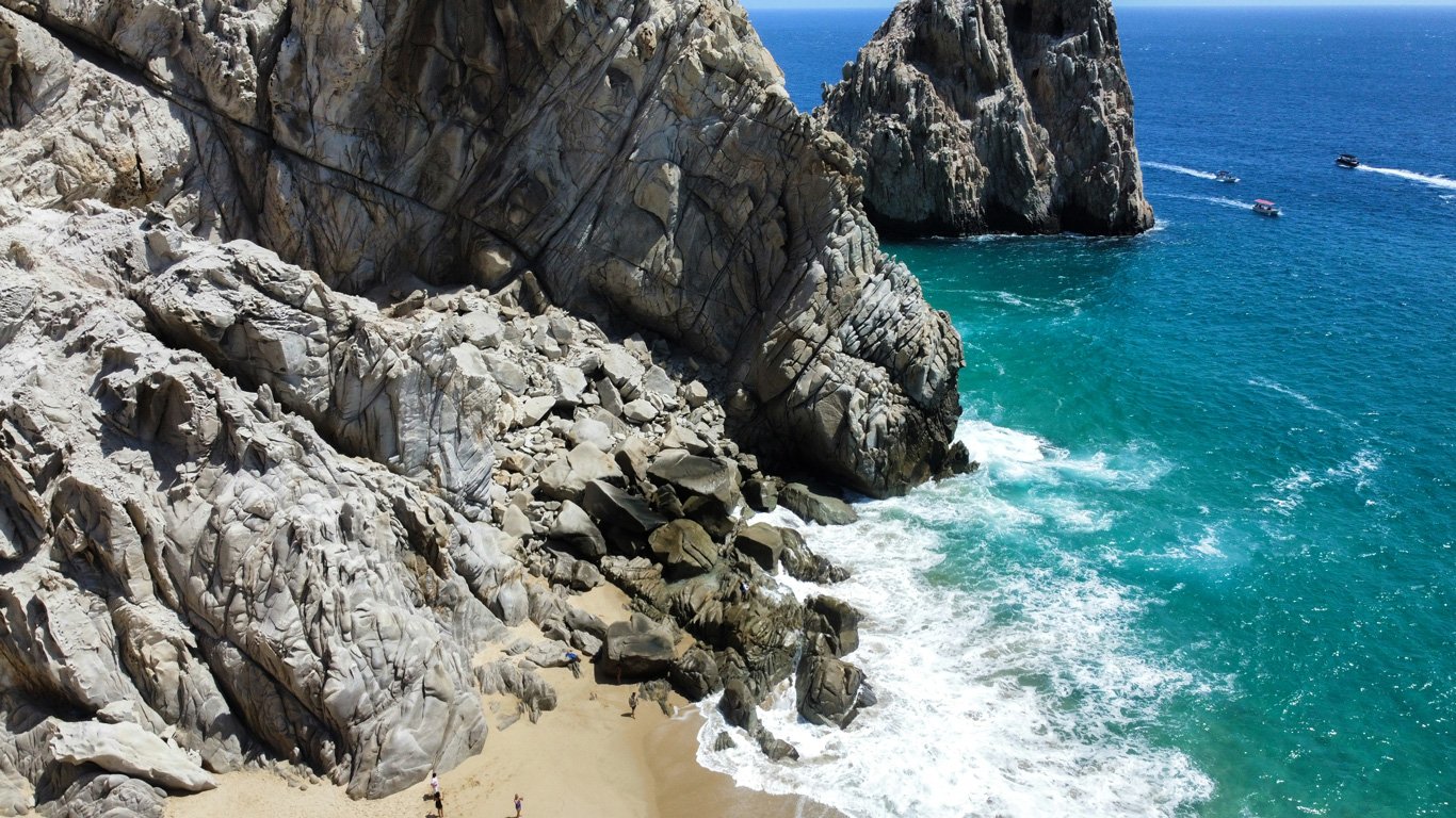 Um litoral dramático com rochas imponentes em Cabo de San Lucas, pequenas praias de areia dourada e ondas azul-esverdeadas quebrando suavemente, criando um refúgio selvagem e cênico.