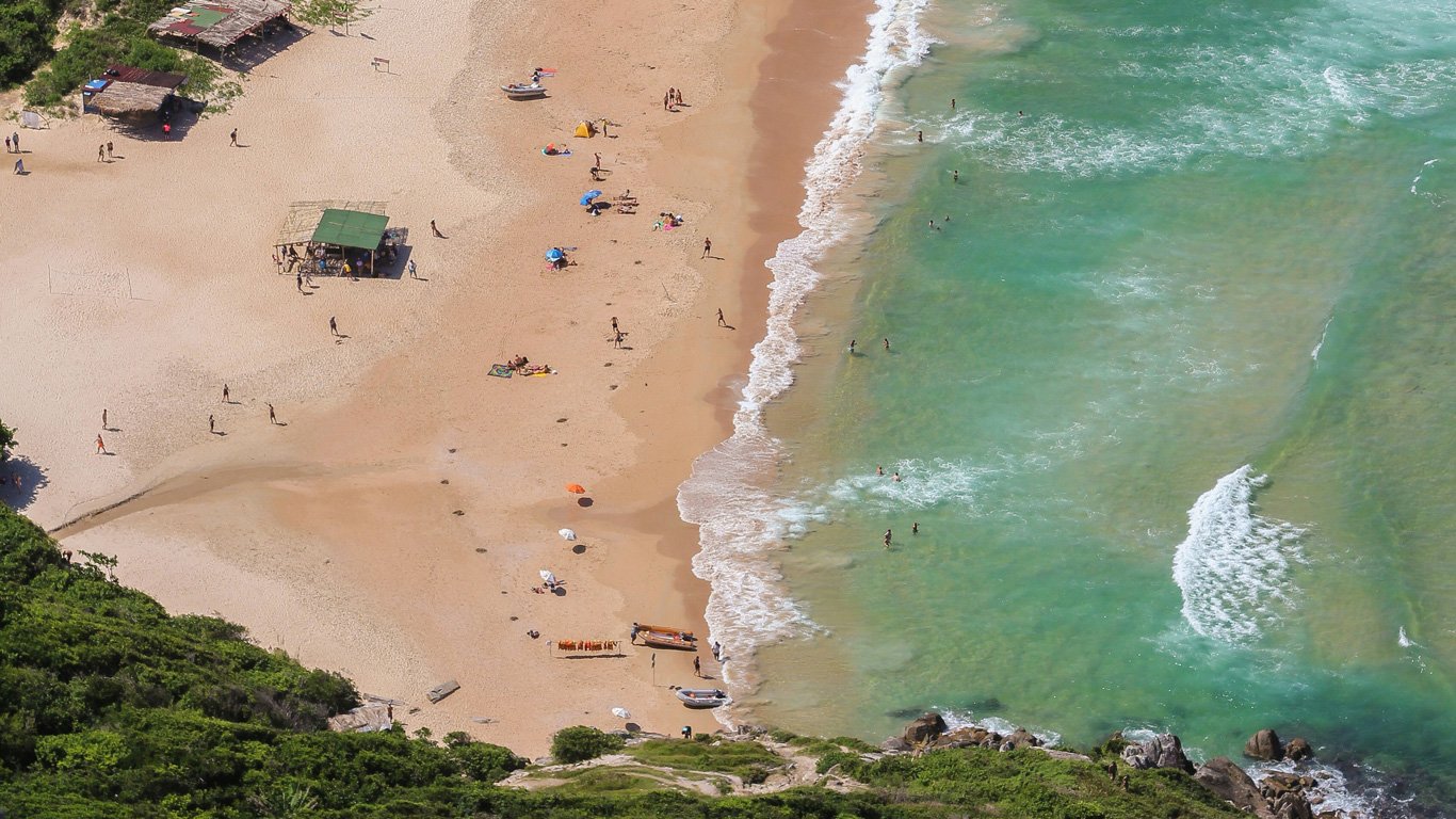 Vista aérea da Praia Lagoinha do Leste em Santa Catarina, com um bar esverdeado com poucas ondas, areias douradas e cercada por vegetação.